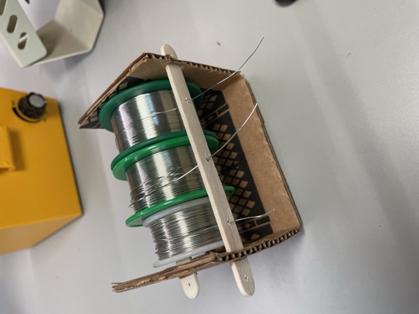 two popsicle sticks shoved through slits in folded cardboard, holding solder spools